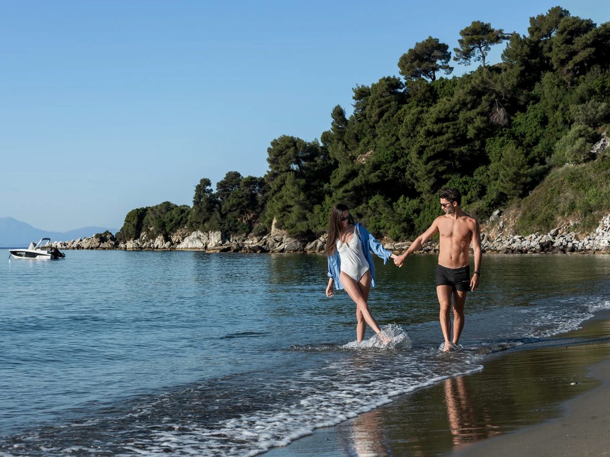 Shallow, calm water over a sandy shoreline sheltered by a rocky, pine-covered headland.