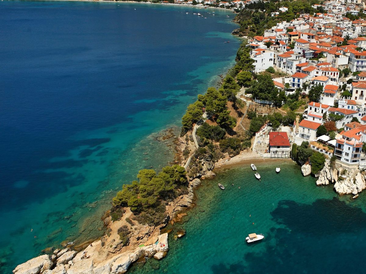 Aerial view of a coastal town of white houses with red roofs above turquoise coves and small boats.