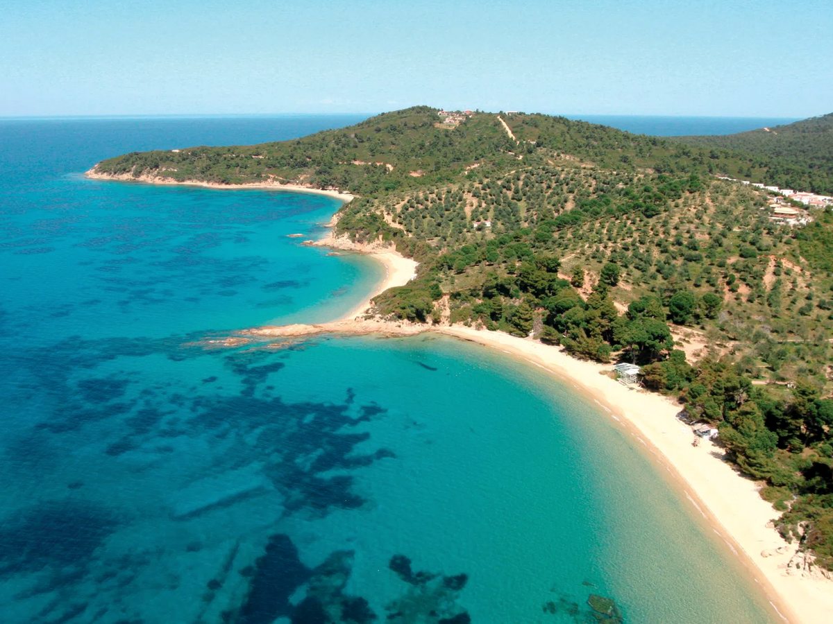 Aerial view of a wide, curving sandy beach along a turquoise bay backed by wooded hills.