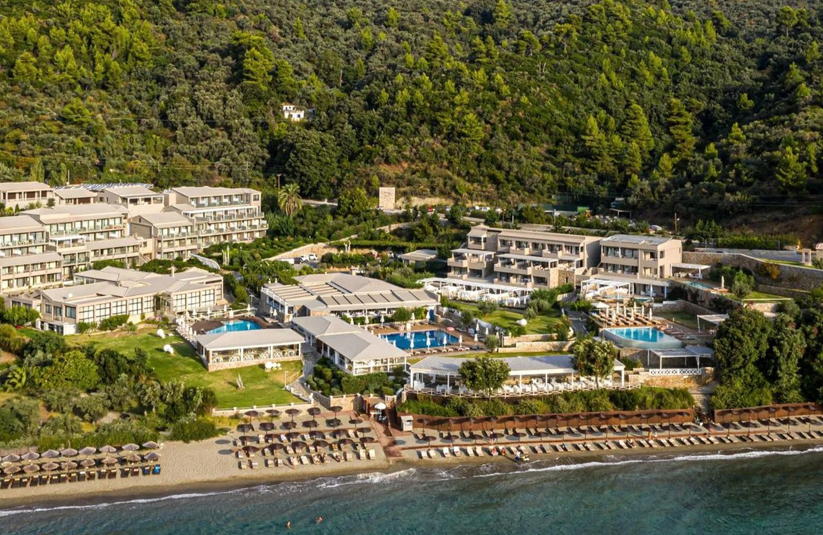 Kassandra Bay Resort from the air: low-rise buildings and pools above a sandy beach lined with straw parasols on Vasilias Bay in Skiathos, backed by a wooded hillside.