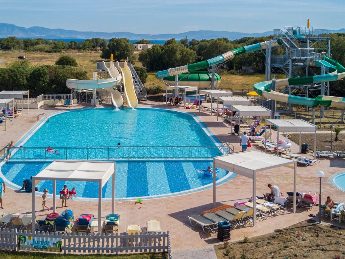 Green and cream water slides on a tall metal frame beside a curved pool, with a fenced junior club area and the sea beyond.