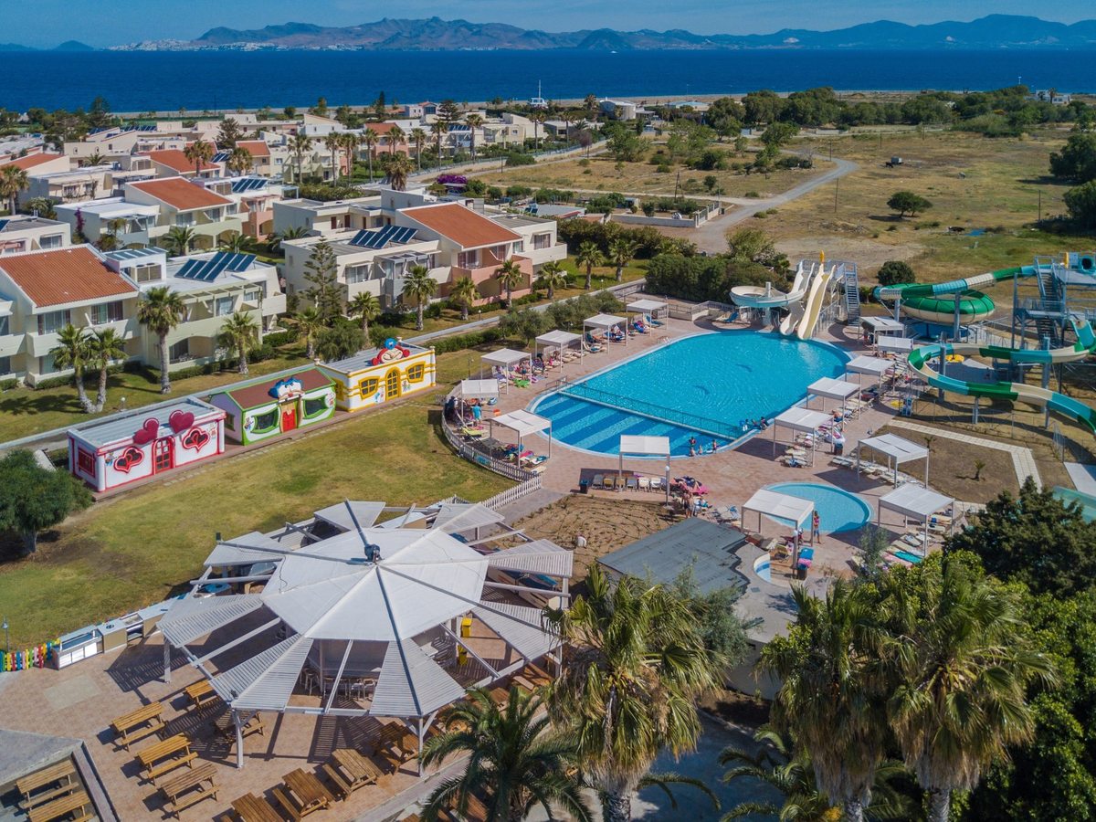 Aerial view of the resort's pools, water slides, colourful club buildings and accommodation blocks, with the sea and mainland hills beyond.