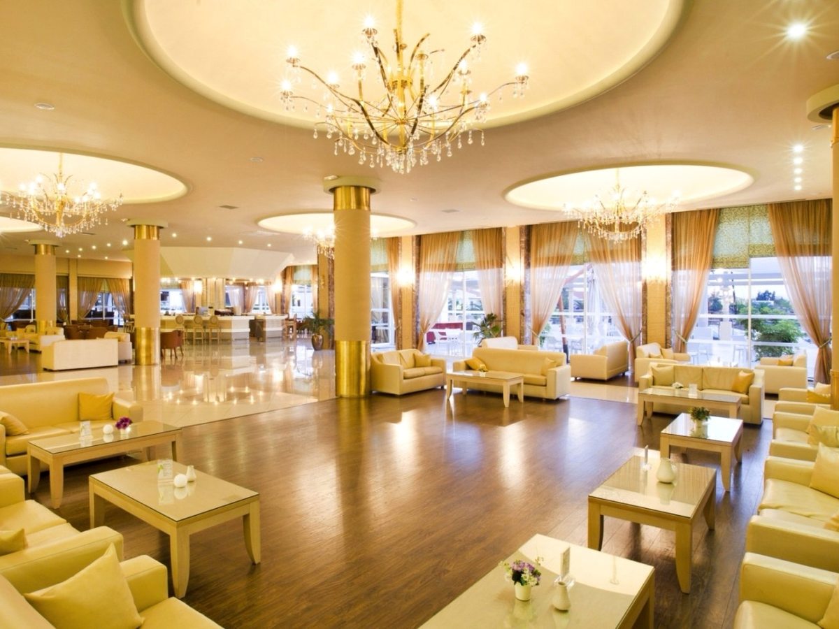 A large lounge with gold chandeliers, cream and yellow sofas and armchairs, marble columns and a polished floor.