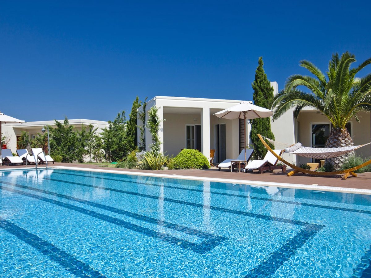 A long pool edged with wooden loungers, white parasols and a hammock beside single-storey bungalow rooms under a clear blue sky.