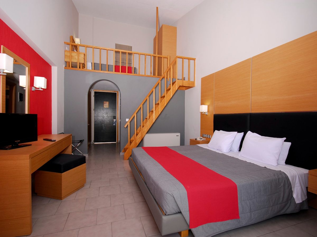 A room with a double bed below and a railed mezzanine above reached by an internal wooden staircase, with a red feature wall.