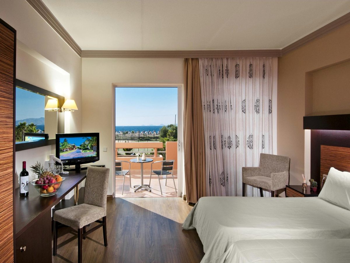 A twin room with a desk, mirror and a welcome fruit bowl, its glass doors open to a balcony table and chairs and the sea and distant hills beyond.