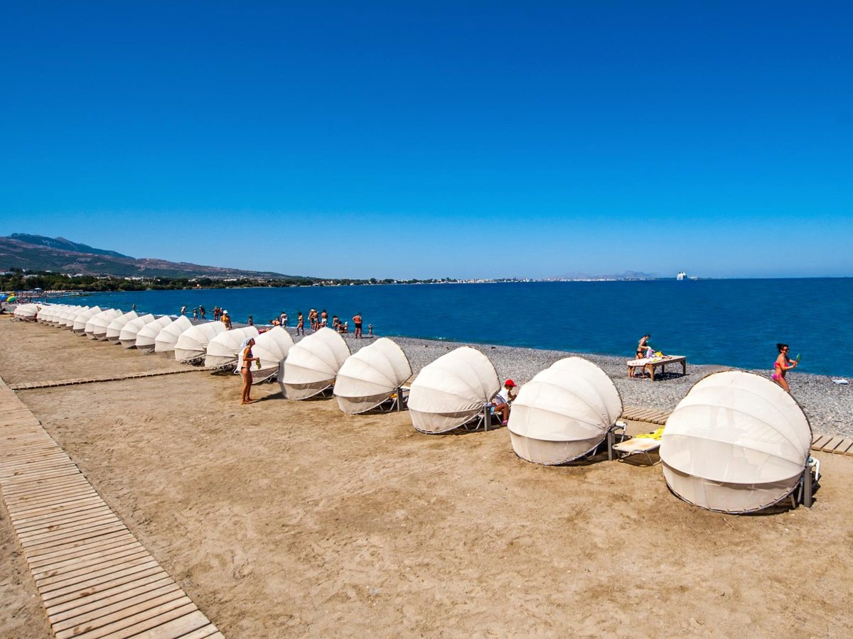 A row of white shell-shaped sun shades along a broad shingle beach beside a calm blue sea, with hills and a town across the bay.