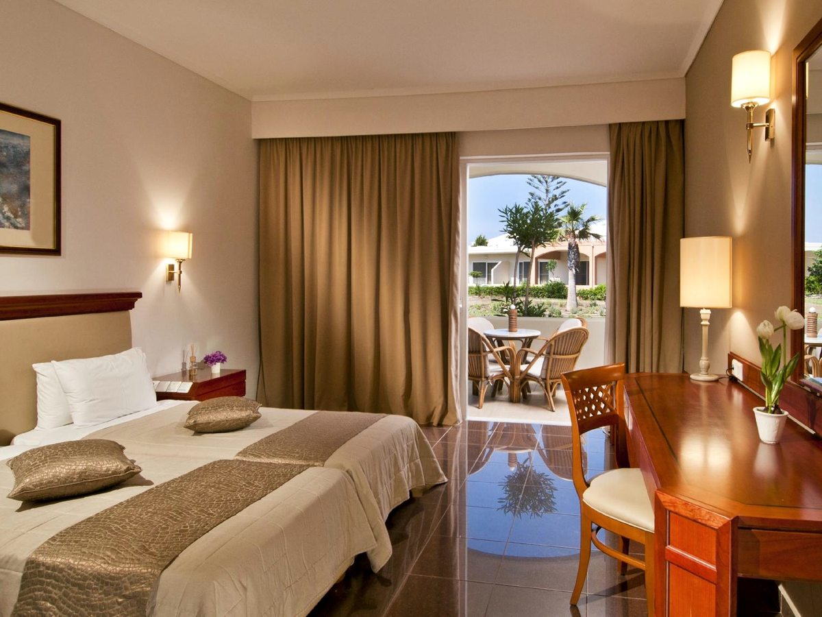 A twin room with cream and gold bedding and a polished wooden desk, its doors open onto a terrace with cane chairs, a table and a garden view.