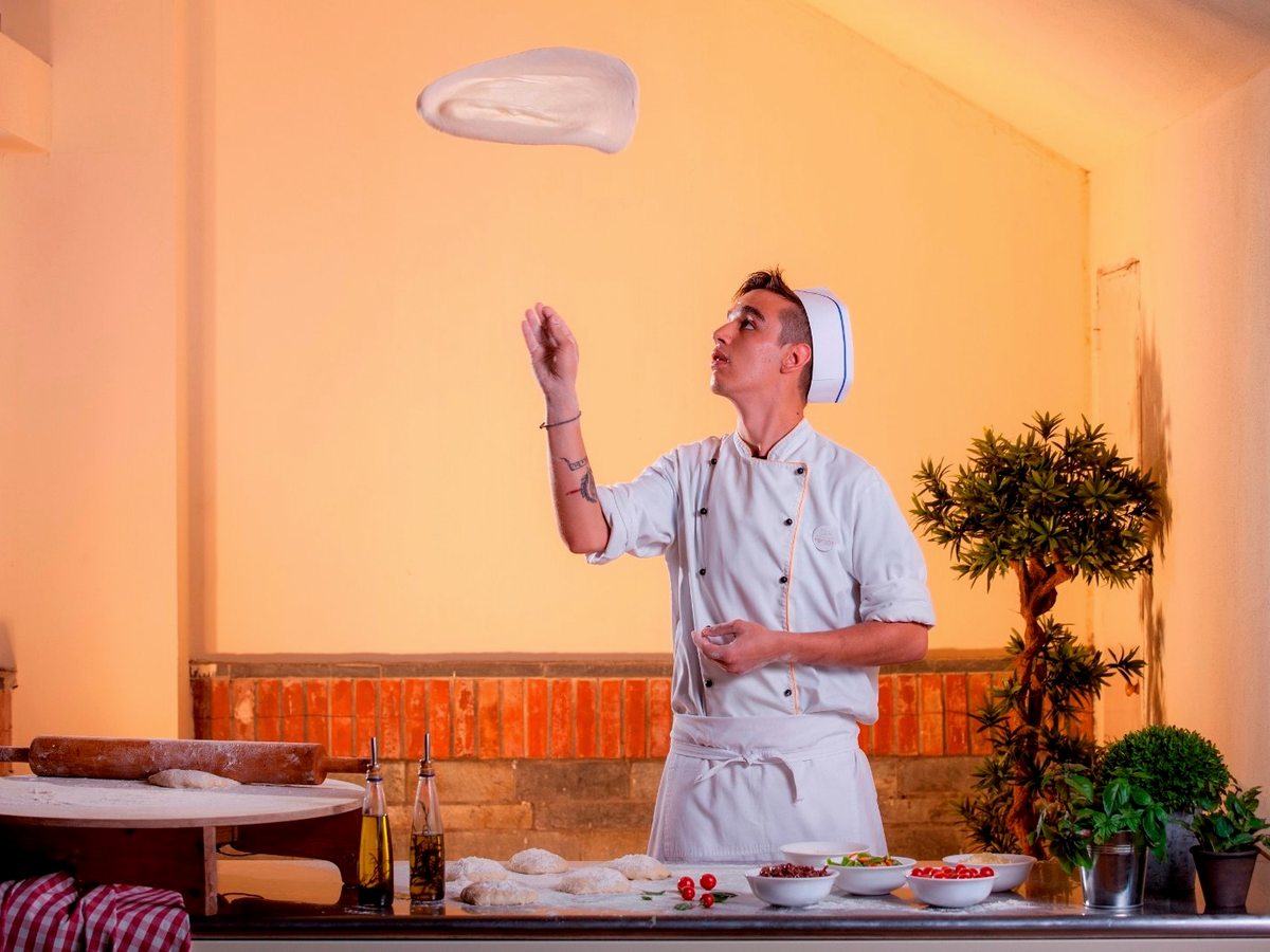 A chef in whites tossing pizza dough at a preparation counter set with flour, olive oil, tomatoes and herbs against a brick and cream wall.
