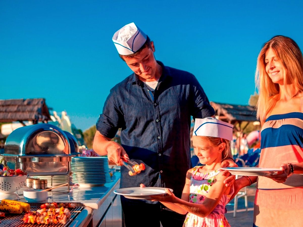 A chef in a paper hat serving grilled meat and corn from a barbecue to a girl and a woman holding plates under a deep blue sky.