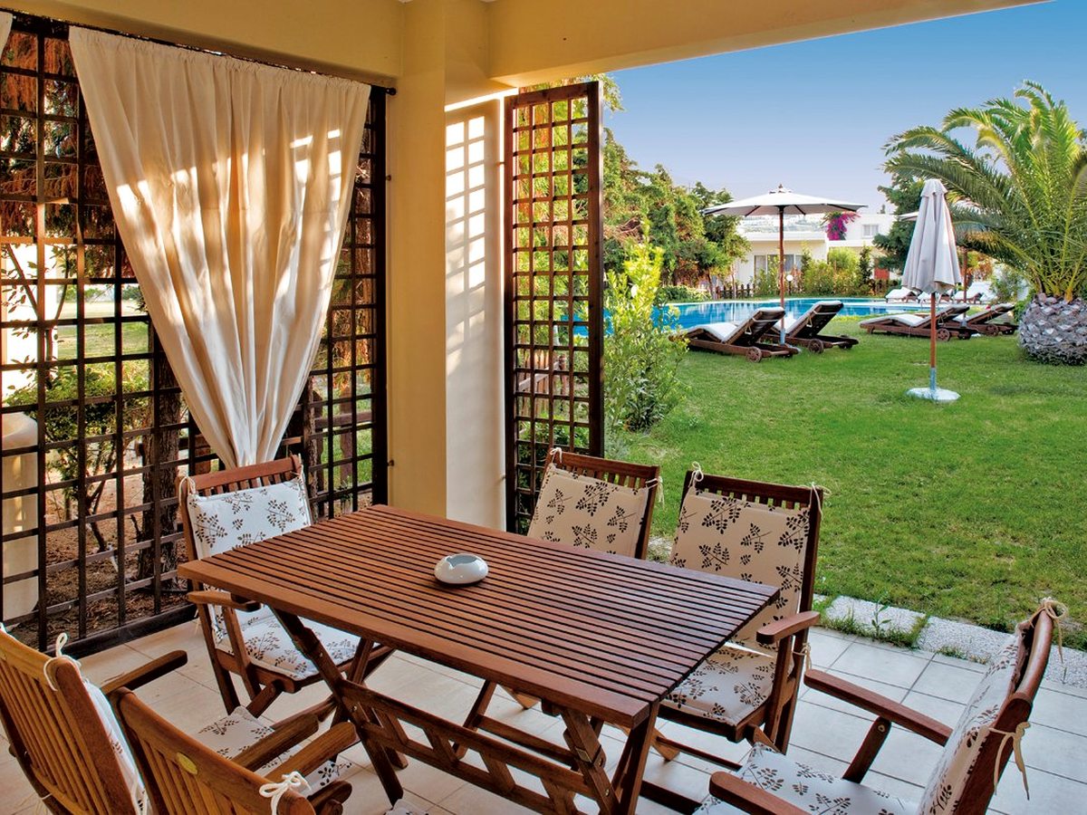 A covered terrace with a wooden table and cushioned chairs opening onto a lawn with sun loungers, a parasol and a pool beyond.