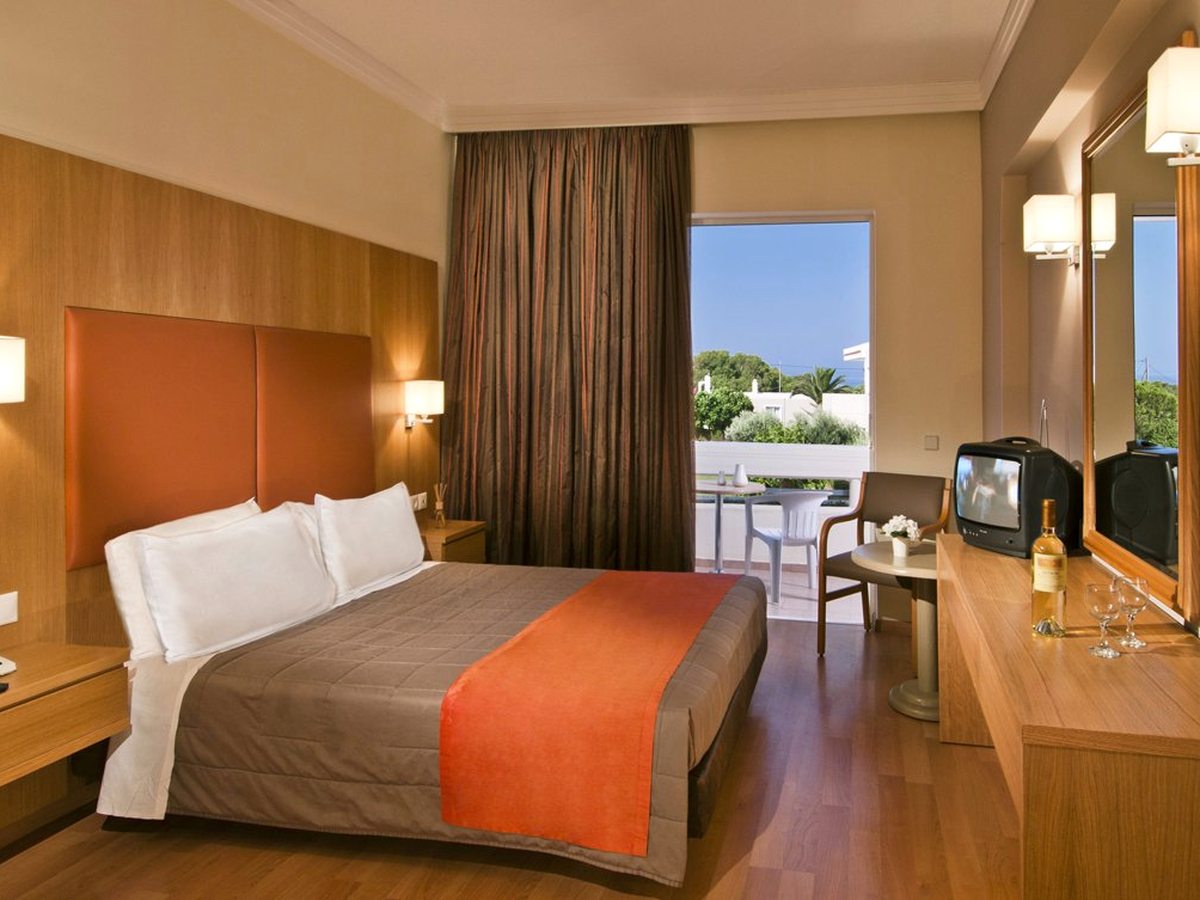 A double room with a wood-panelled headboard, an orange bed runner, a desk and TV and glass doors to a small furnished balcony with a garden view.