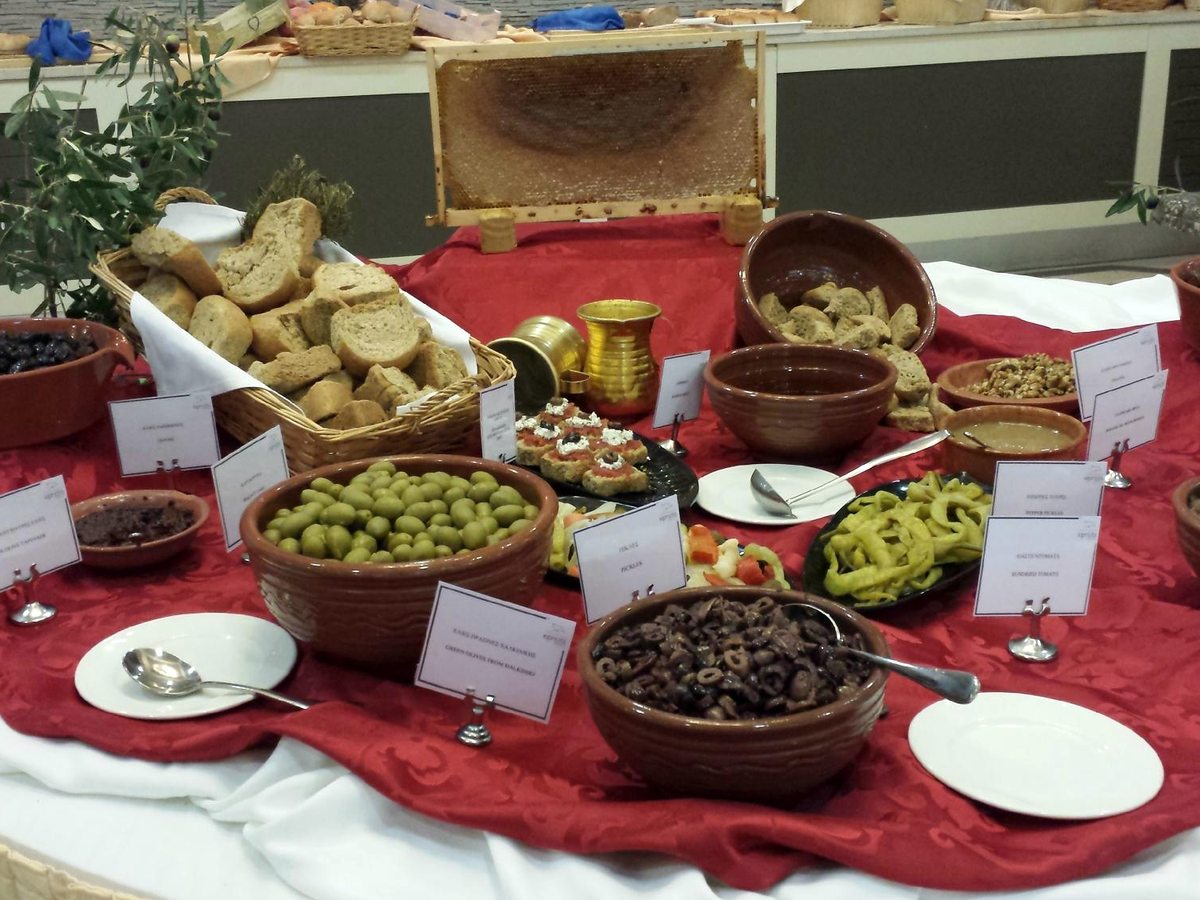 A buffet table with a red cloth laid with bowls of green and black olives, breads, dips and pickled peppers, each dish labelled.