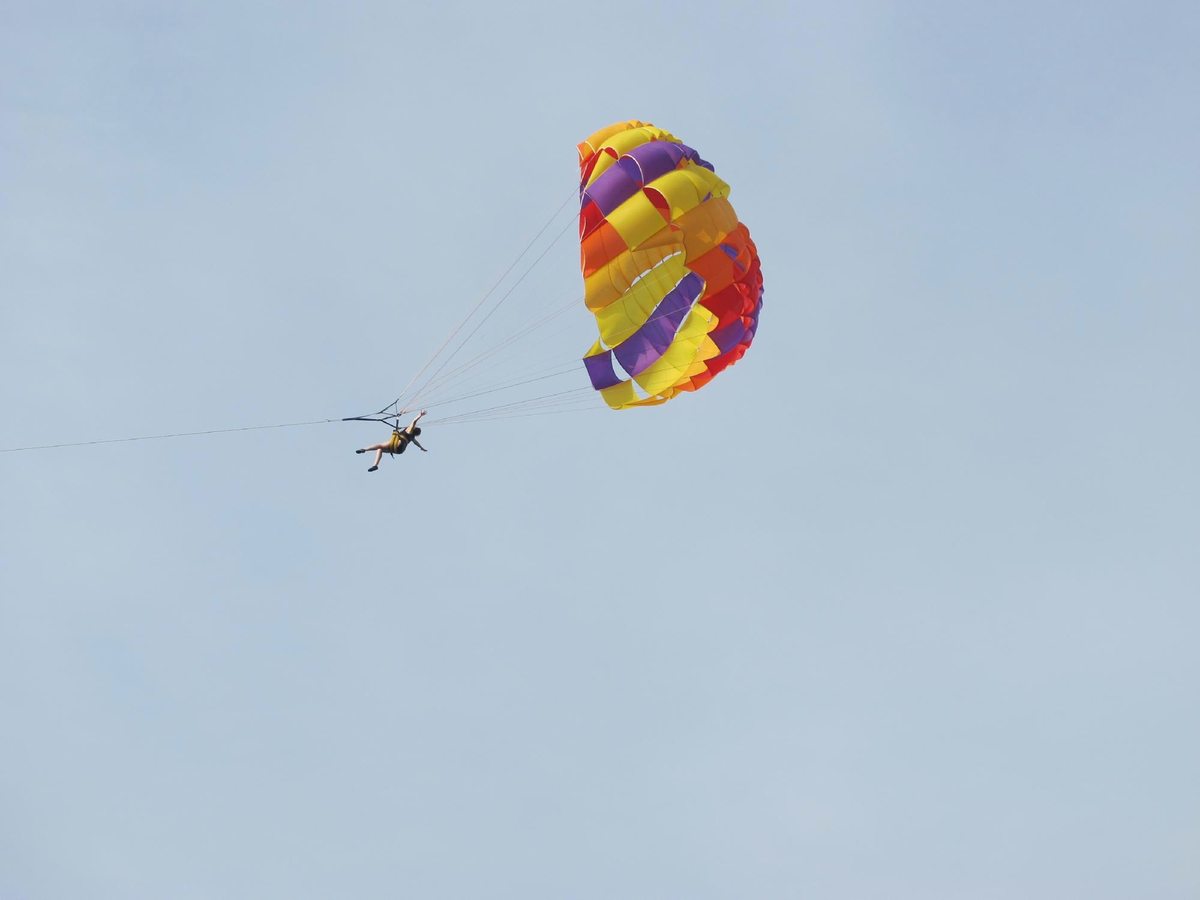 A parasailer in a harness lifted under a red, yellow and purple parachute against a pale blue sky.