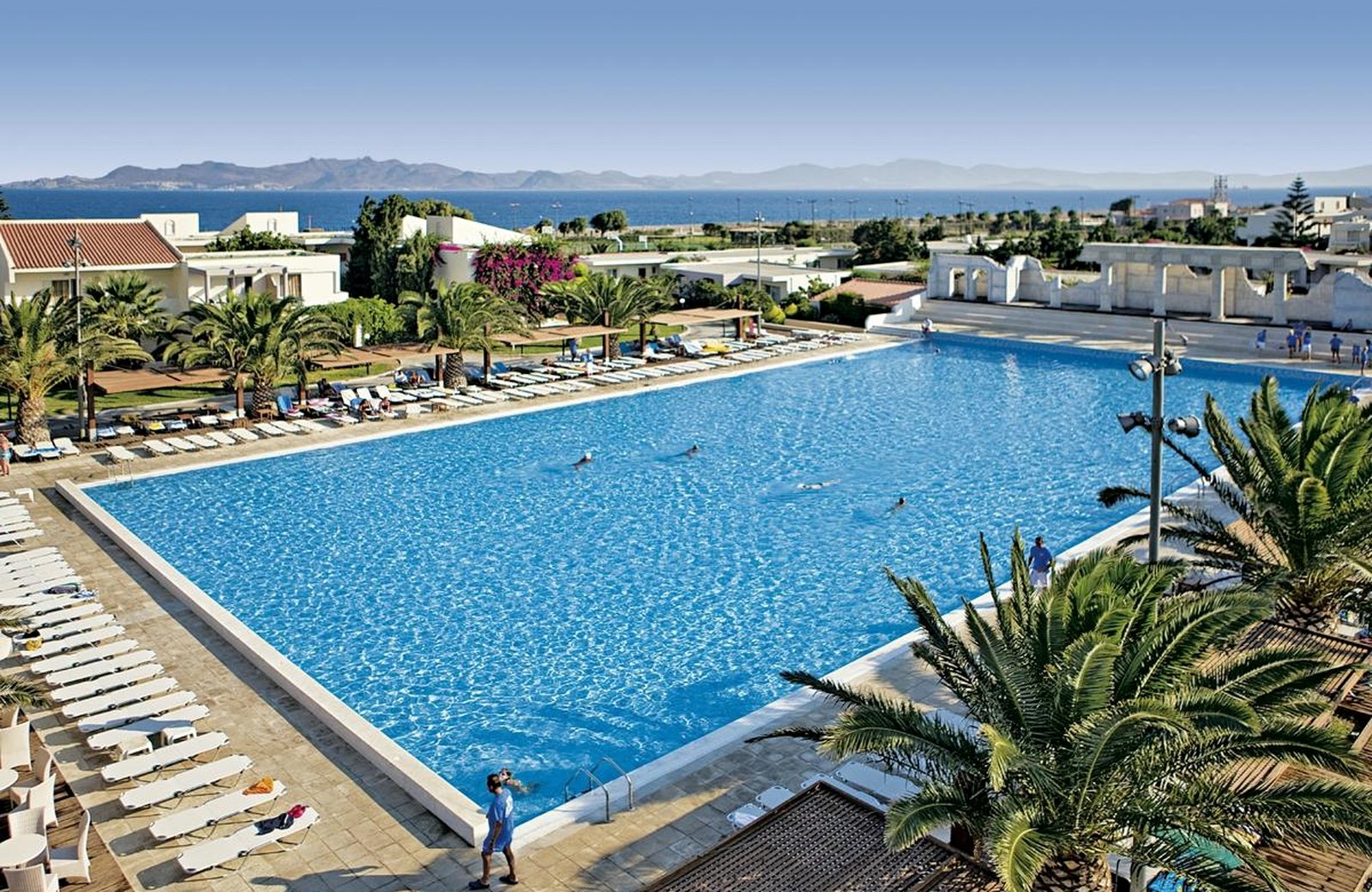 The main pool at Kipriotis Village Resort, a large rectangular pool edged with white sun loungers and palms, with the Aegean and distant hills beyond.