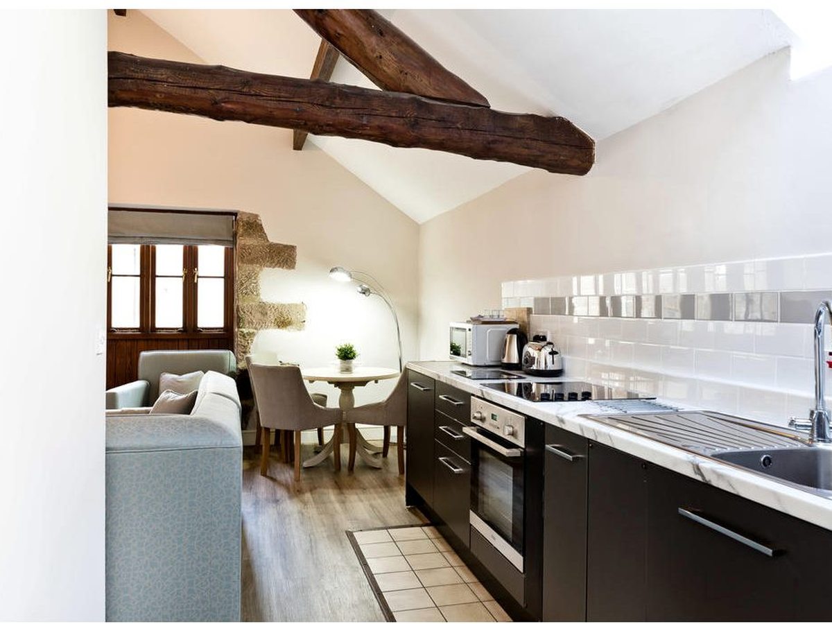 A narrow galley kitchen with dark units, a marble-effect worktop, a built-in oven, hob and microwave under a sloping ceiling with a heavy exposed beam, and a small round dining table beyond.