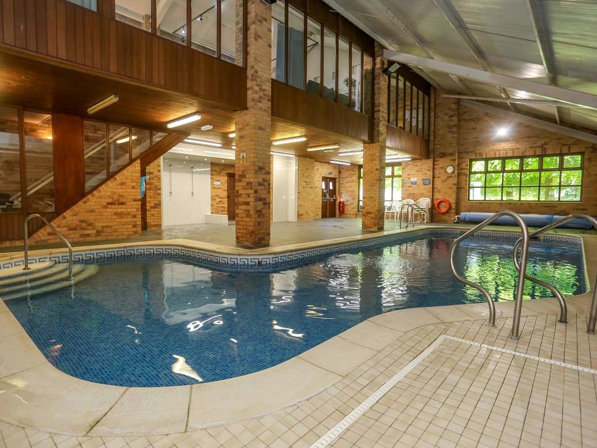 An indoor swimming pool with a curved shallow end and steps into the water, a mosaic tile border, brick pillars and a timber gallery above, and windows onto trees.