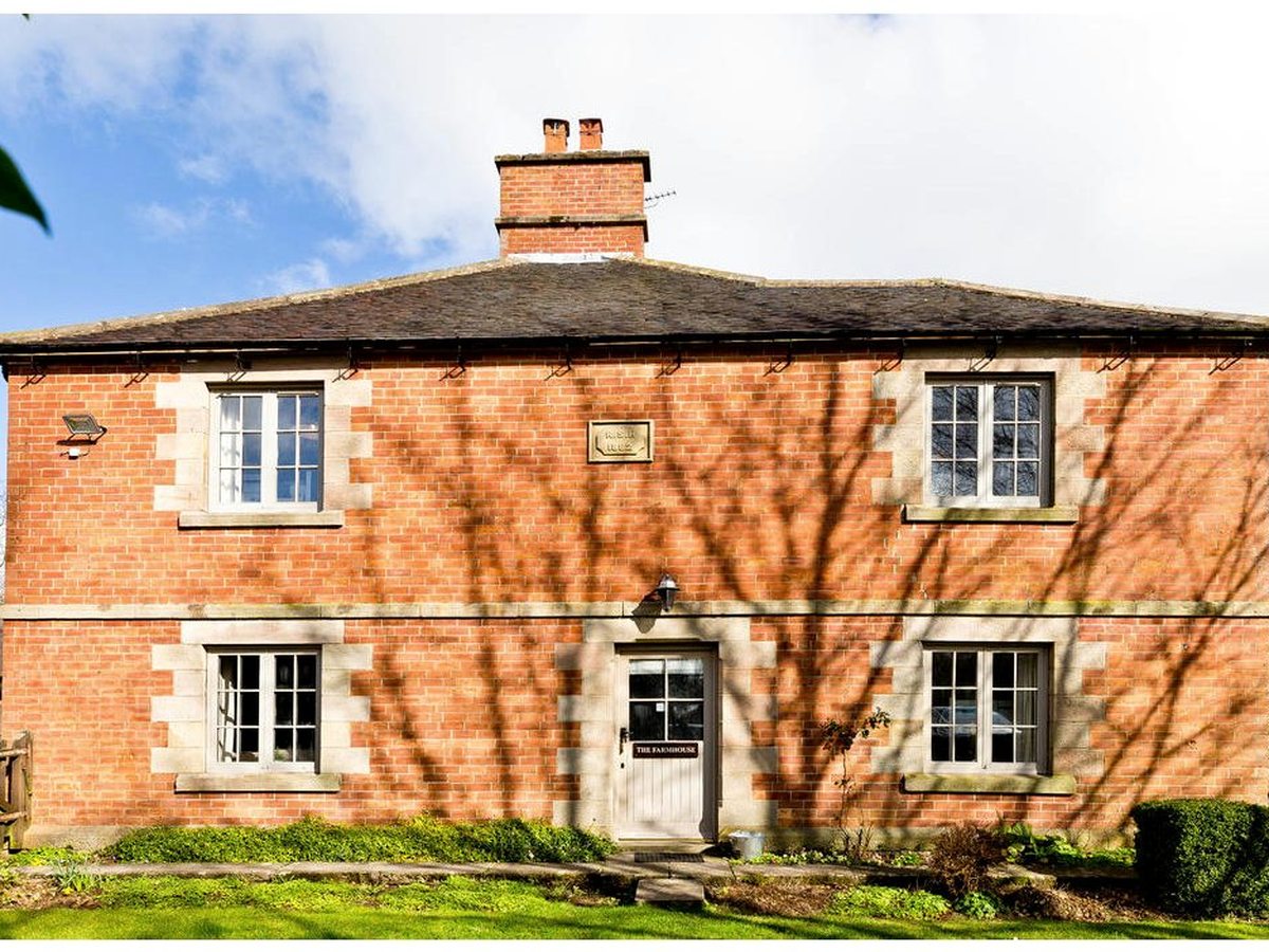 A red-brick two-storey farmhouse with pale stone quoins, white-framed windows, a central front door with a nameplate and a lawn and low border in front.