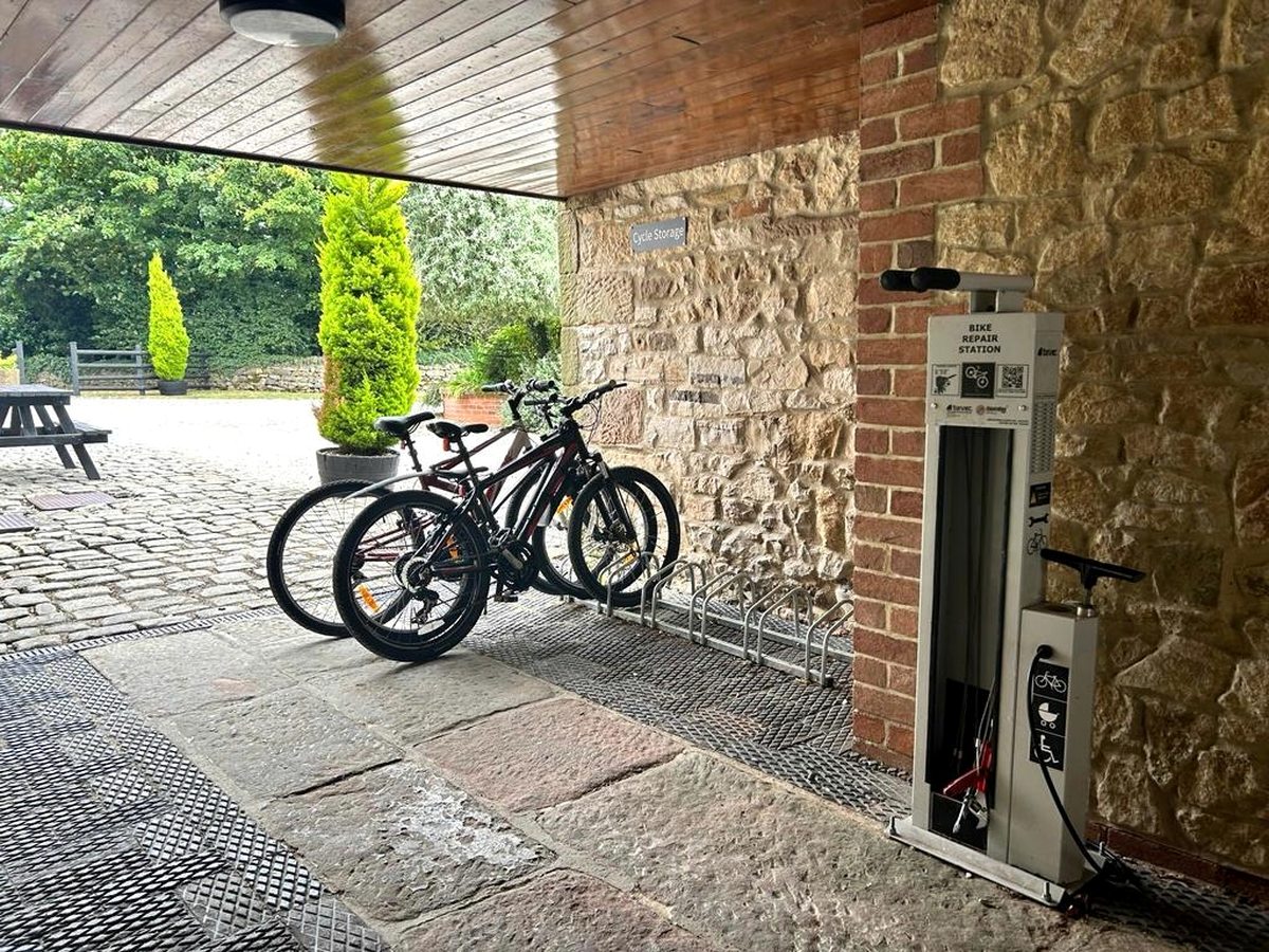 Bicycles in a metal rack under a covered stone archway, with a wall sign reading Cycle Storage and a bike repair station and floor pump alongside.