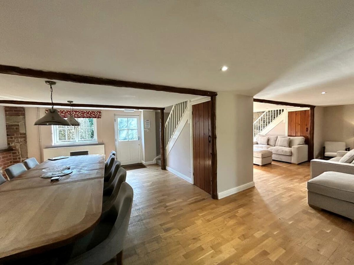 A large open-plan cottage interior with a long wooden dining table and upholstered chairs under pendant lights, a wood floor, exposed beams and a sitting area with cream sofas beyond.