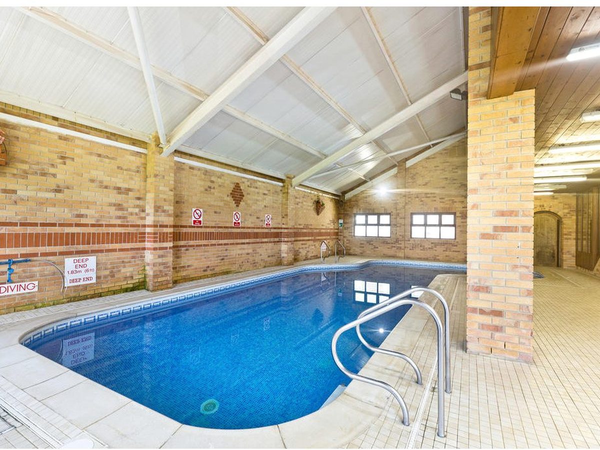 The far end of the indoor pool under a white pitched roof, with brick walls, a handrail into the water and red and white safety signs including a deep-end depth notice.