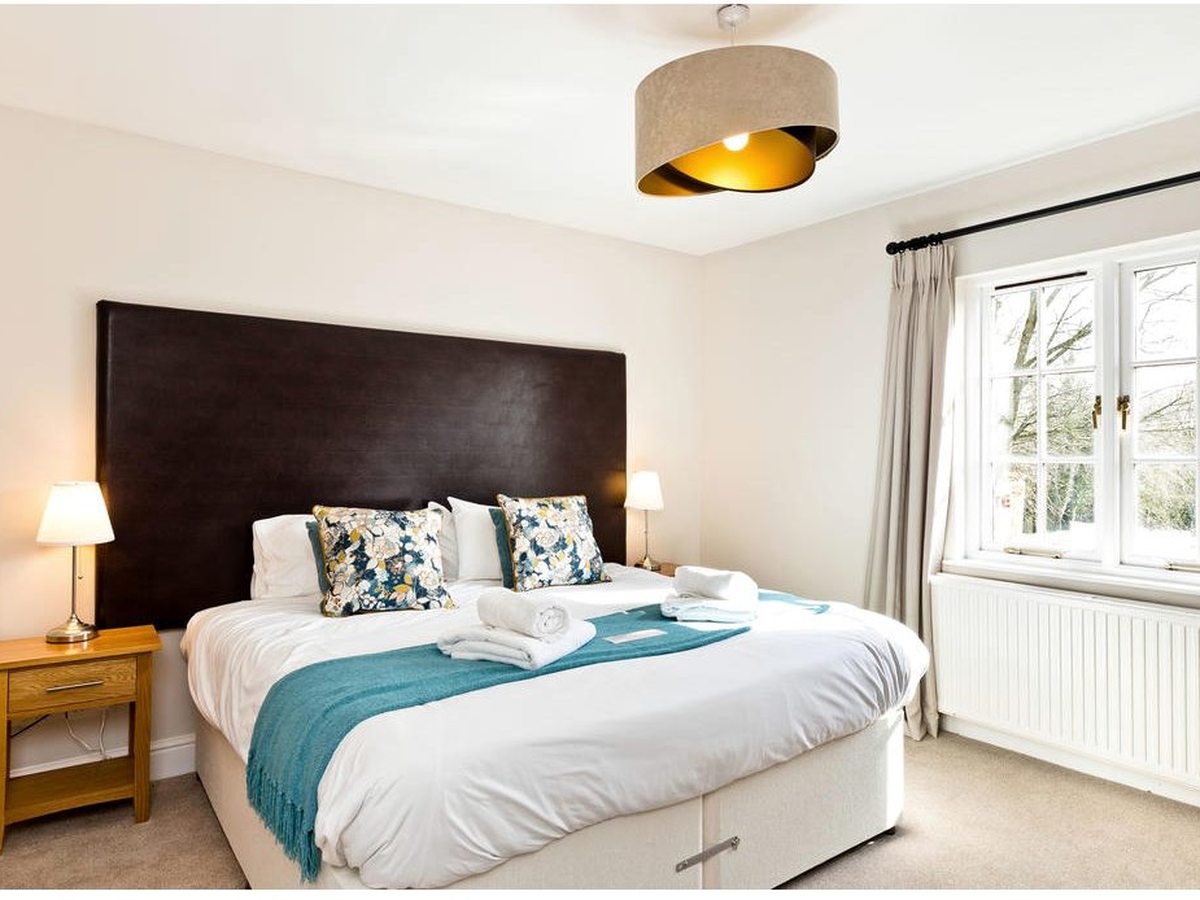 A bright double bedroom with a tall dark leather headboard, white bedding with a teal throw and folded towels, bedside tables with lamps and a window onto trees.