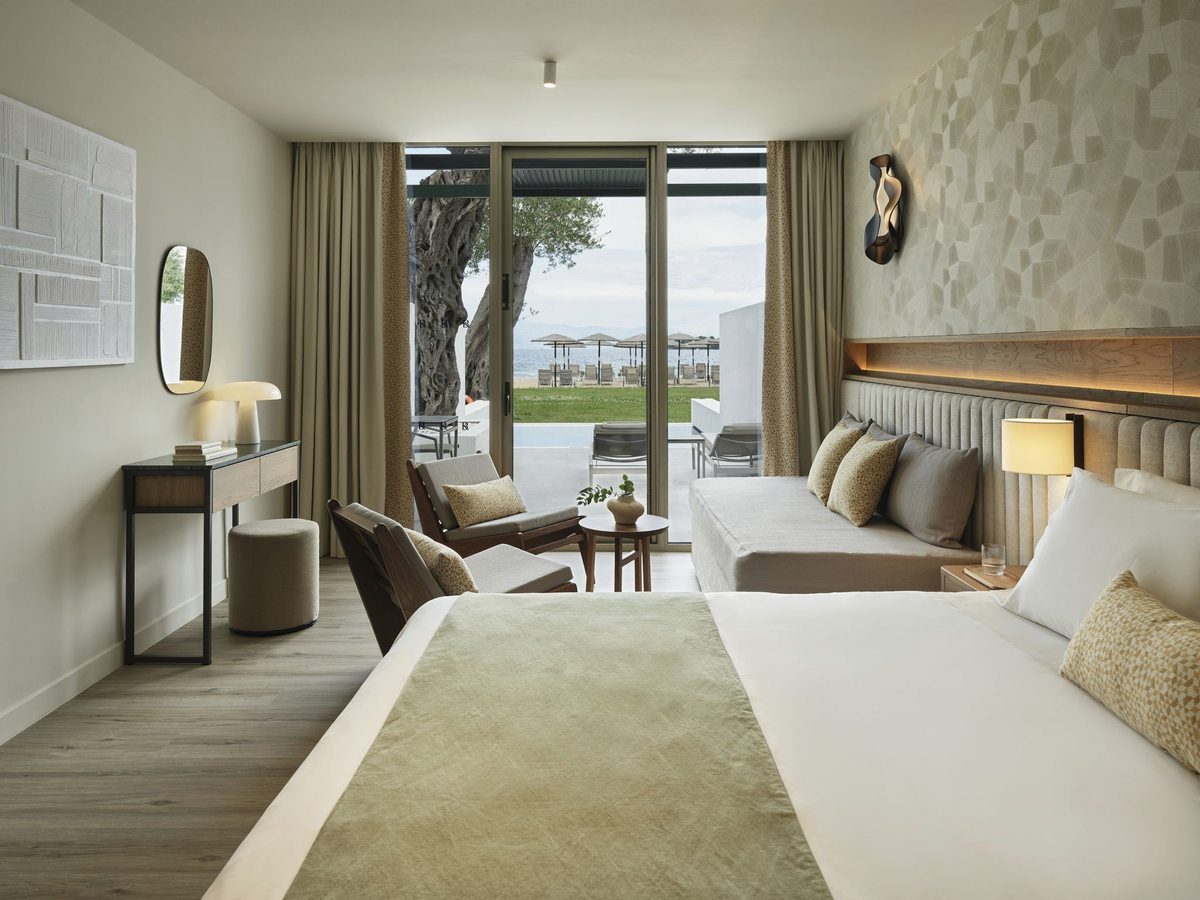 A bedroom with a wide bed, a long sofa and sliding doors open to a paved terrace, with lawn, an olive tree and straw beach umbrellas beyond.