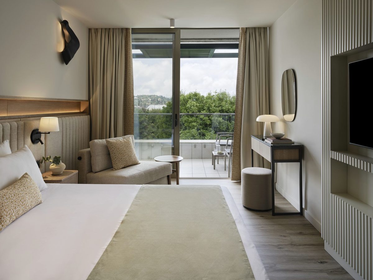 A bedroom with a wide bed, a small sofa and sliding doors to a balcony with a table and chairs, greenery and a green hillside beyond.