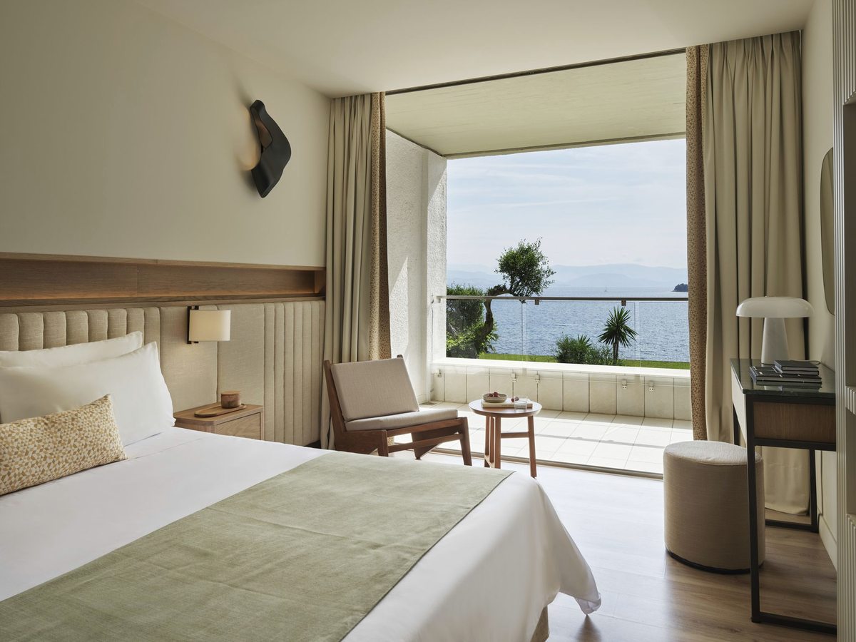 A bedroom with a wide bed and a wooden armchair beside a deep open balcony with a glass balustrade, the sea and mountains beyond.