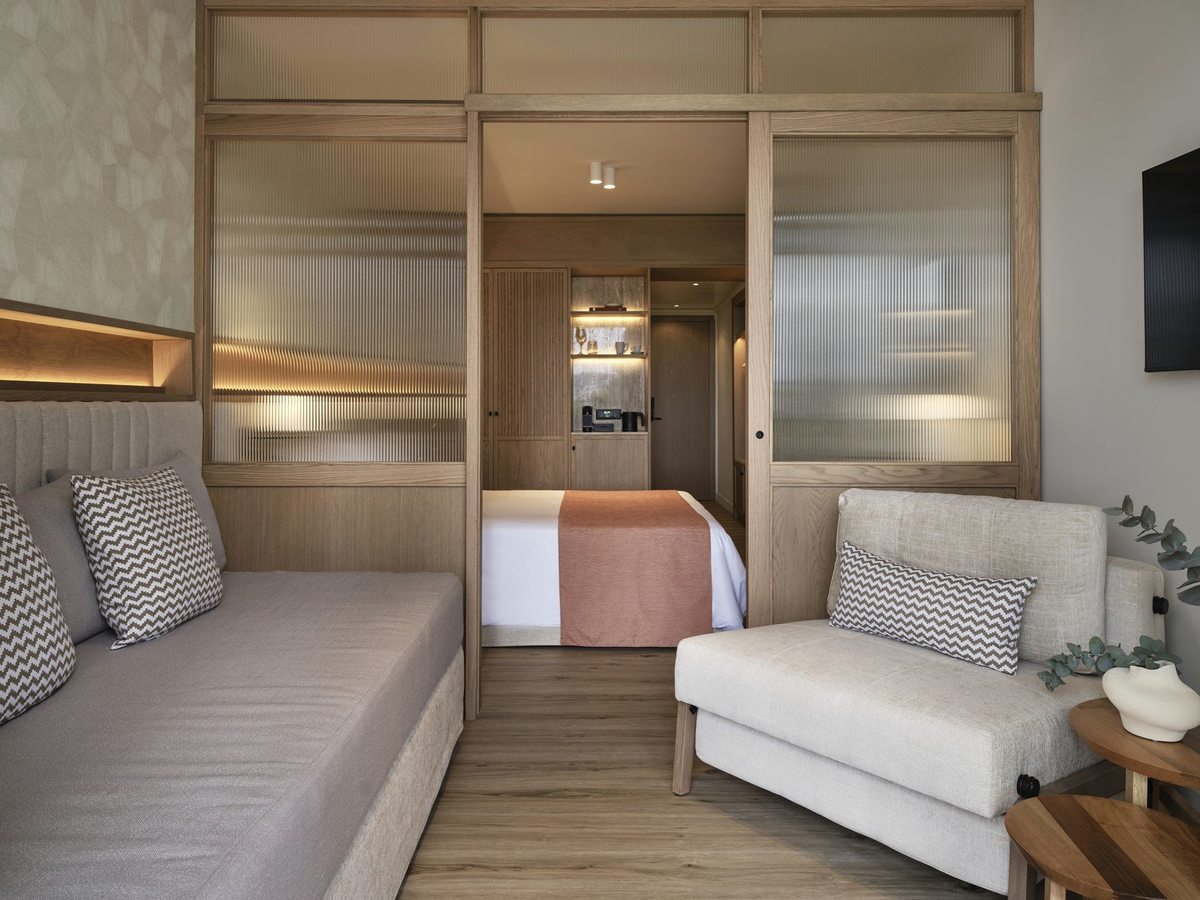 A suite sitting area with a grey sofa bed and a cream armchair, the double bed visible through a wide sliding partition of fluted glass and pale oak.