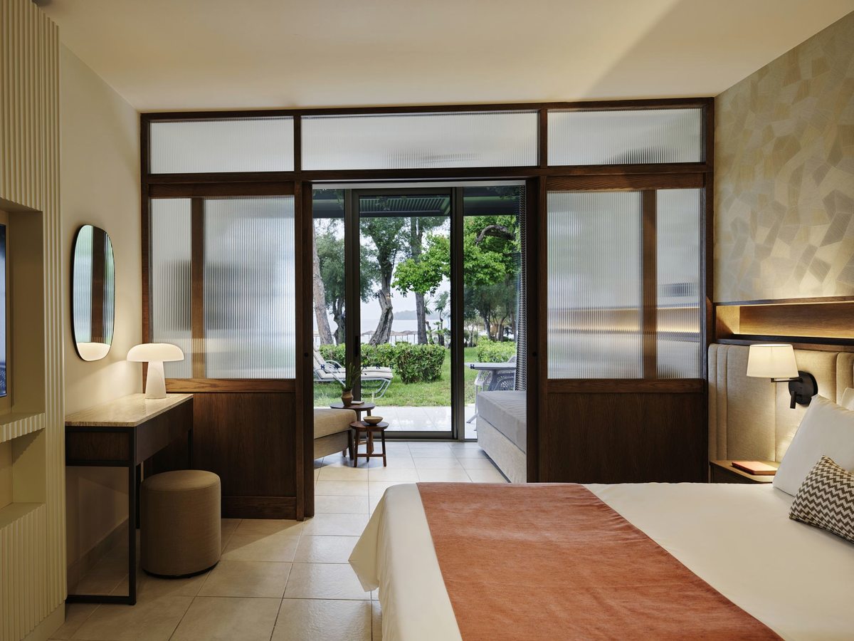A bedroom with an orange throw, divided from a sitting area by a sliding partition of fluted glass in dark wood, with a terrace and pine trees beyond.