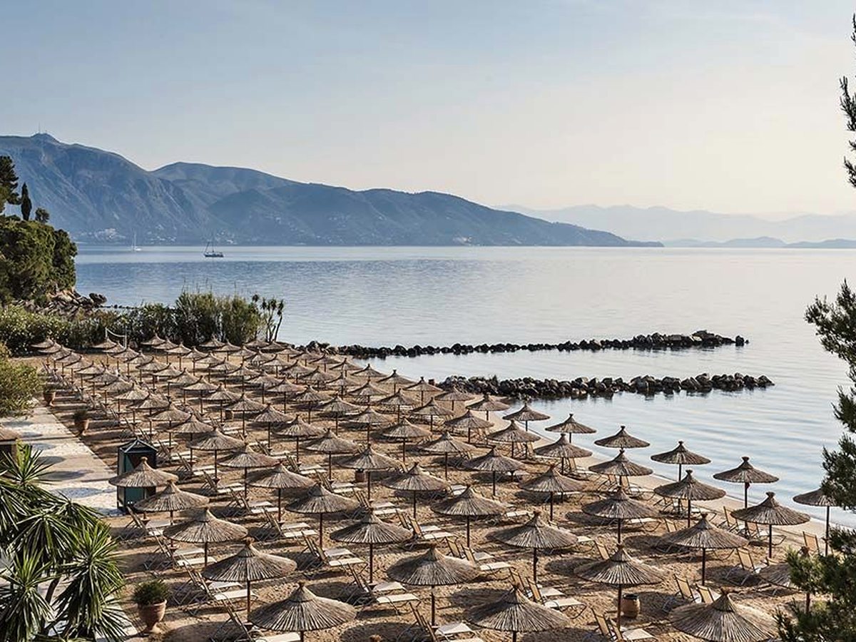 Rows of straw parasols and loungers on a sandy beach beside flat calm water, with a low stone breakwater and mountains across the water.