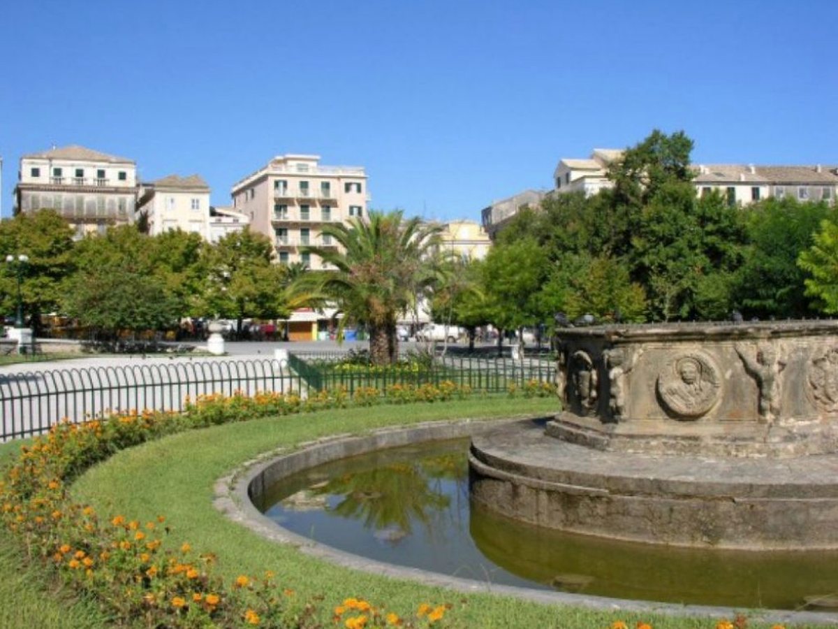 A carved stone fountain in a public garden with marigold beds, a palm tree and pastel Venetian-style townhouses behind.