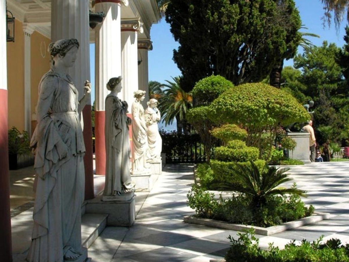 Marble statues of the Muses along a columned terrace at the Achillion Palace, with clipped shrubs, palms and cypresses in the formal garden.