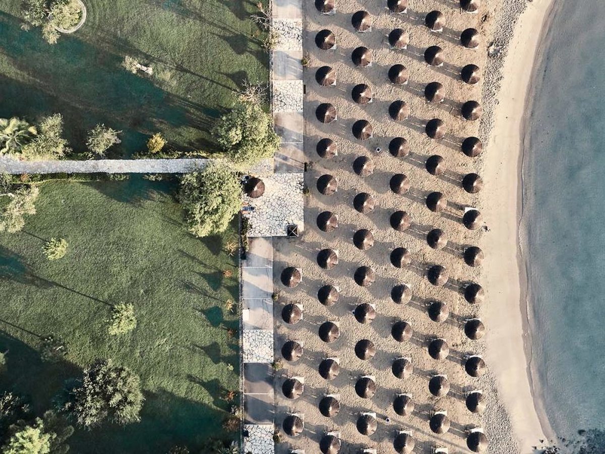 An overhead view of mown lawns, paths and trees running down to rows of straw beach umbrellas on the sand.