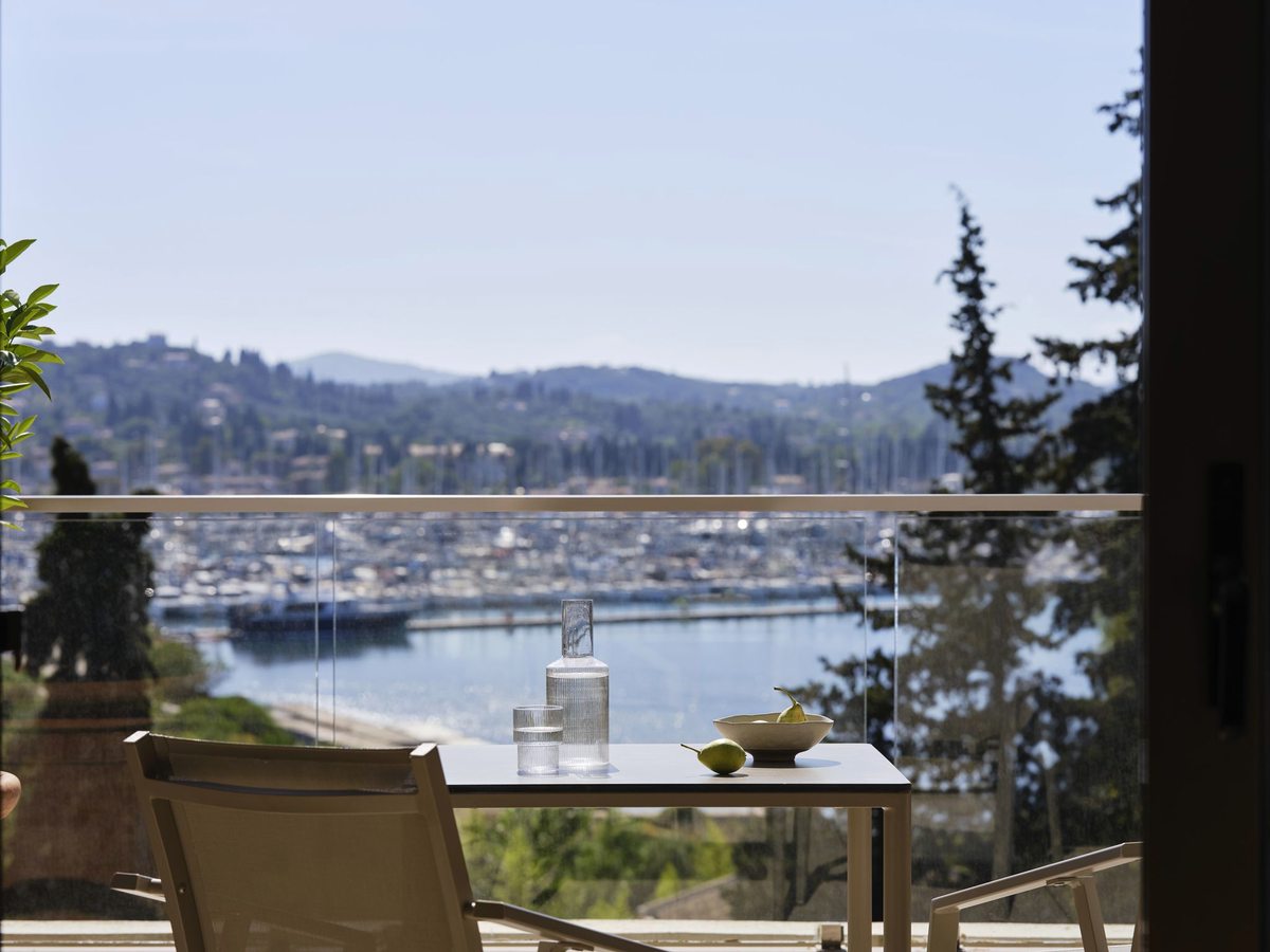 A balcony table and chairs with a water carafe and a bowl of pears, above a marina full of masts with wooded hills beyond.