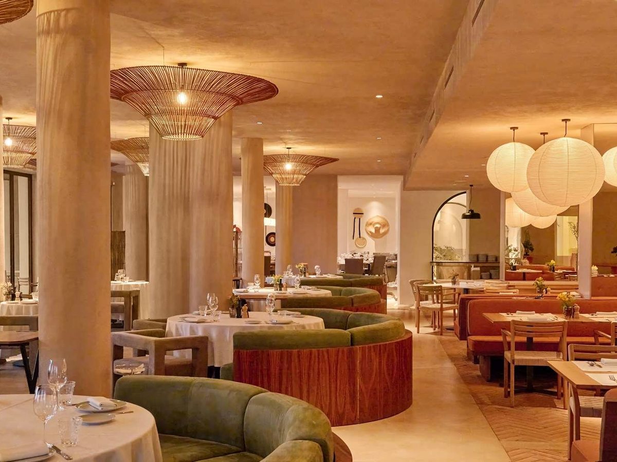 A large restaurant interior with plaster columns, curved green velvet banquettes, white-clothed tables and woven pendant lights.