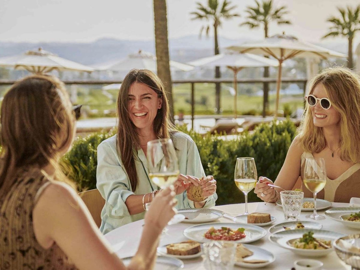 Three women eating and drinking white wine at a round table on a sunlit terrace, with parasols, palms and a golf course behind them.