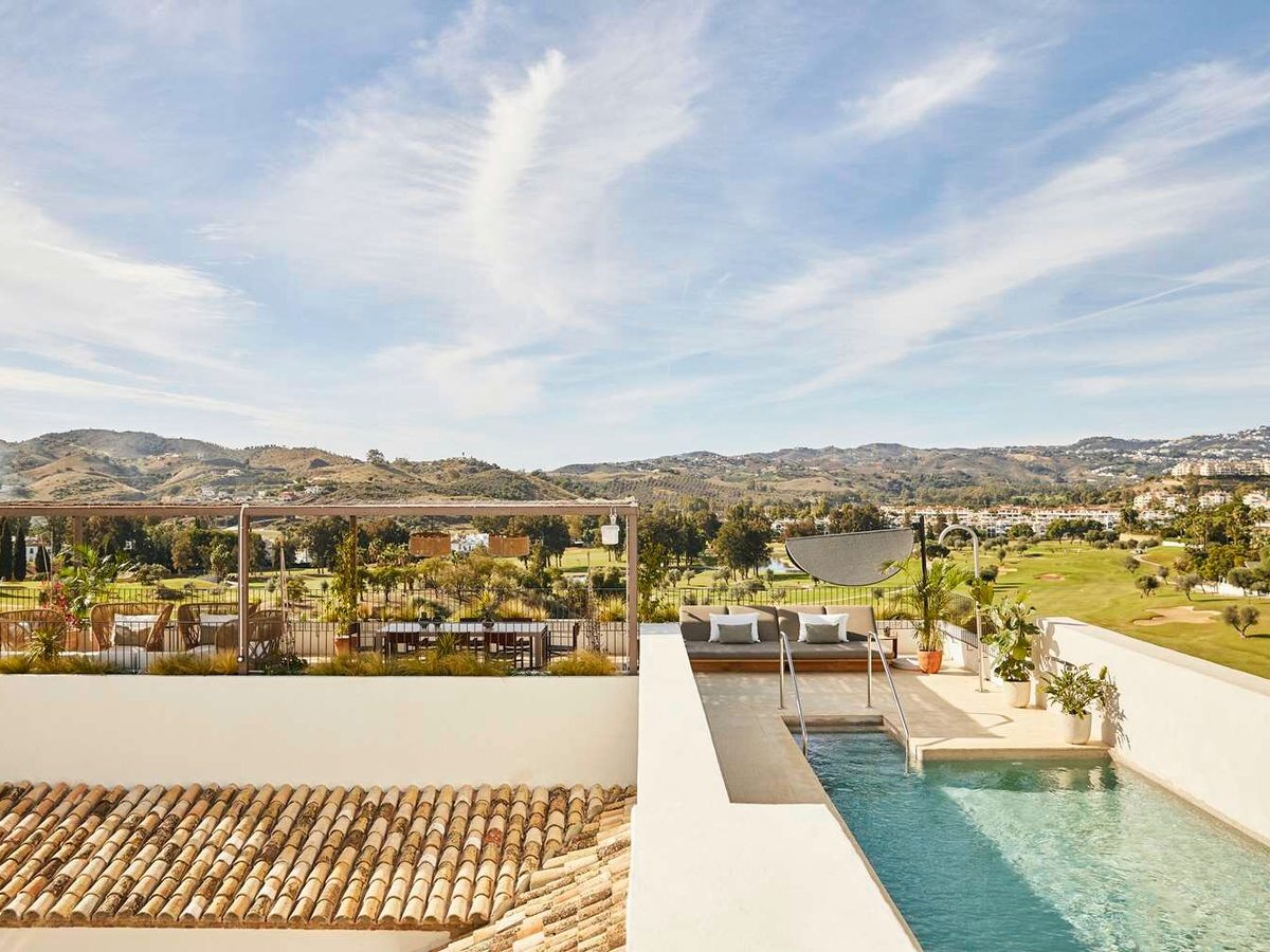 A narrow pool on a private terrace with a cushioned day bed and parasol, above tiled roofs and a golf course with hills behind.