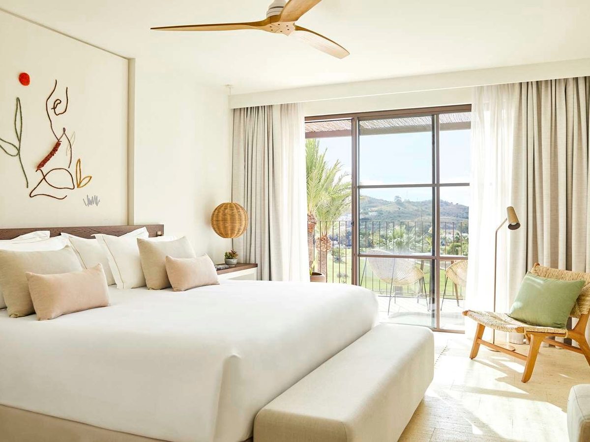 A bright bedroom with a king bed in white linen, a wooden ceiling fan, wire wall art and sliding doors onto a balcony with wicker chairs.