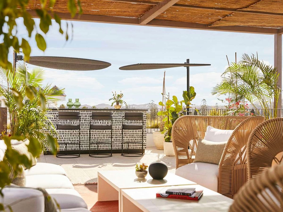 A shaded outdoor terrace with a bar counter and stools, wicker armchairs, low white sofas, potted palms and hills in the distance.