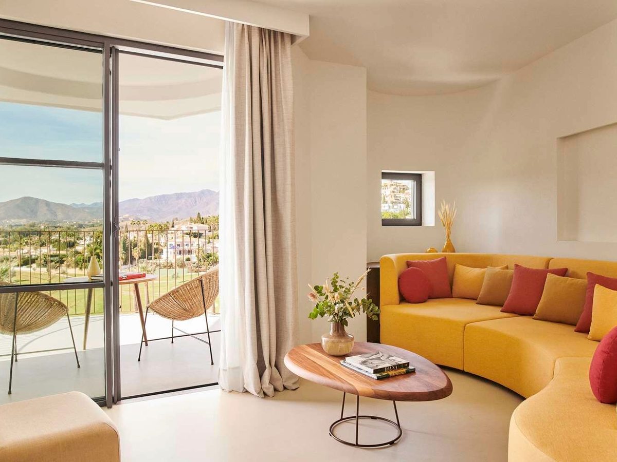 A suite sitting room with a curved mustard yellow sofa, red cushions, an oval wooden coffee table and doors to a balcony with mountains beyond.