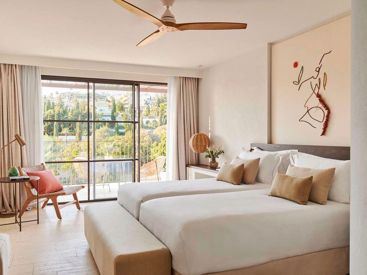 A bedroom with two single beds side by side, cream and mustard cushions, a ceiling fan and sliding doors to a balcony above gardens.