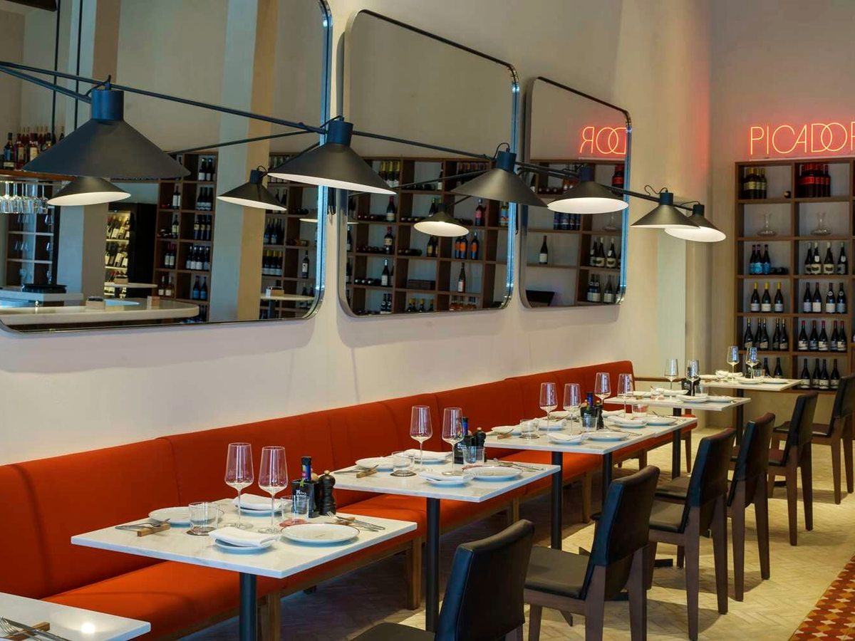 A narrow restaurant with a long orange banquette, white marble tables set with wine glasses, wine racks along the wall and a neon Picador sign.