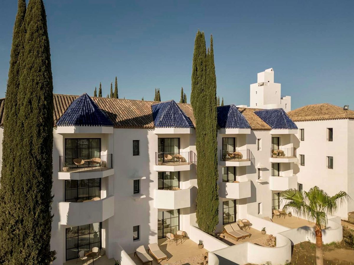White hotel buildings with blue-tiled pitched roofs and curved balconies set with wicker chairs, tall cypress trees in front and a clear blue sky.