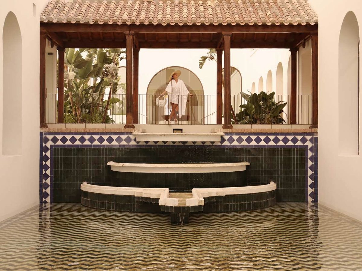 A blue and white tiled water basin with stepped cascades, below a timber pergola where a woman in a robe and sun hat is walking.