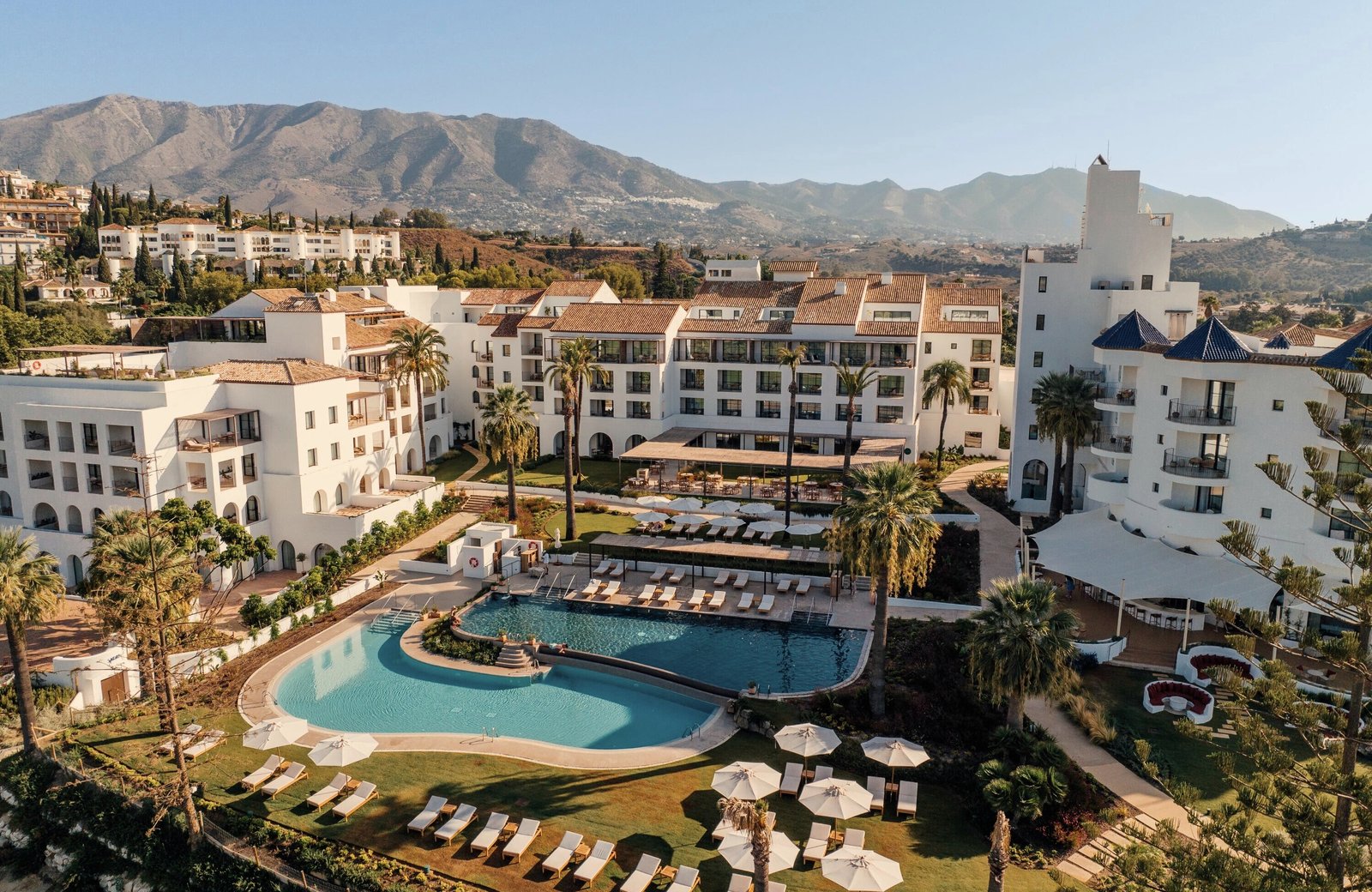 La Zambra Resort from above: white buildings with terracotta and blue-tiled roofs around a large curved pool, lawns, parasols and palms, with the Mijas hills behind.