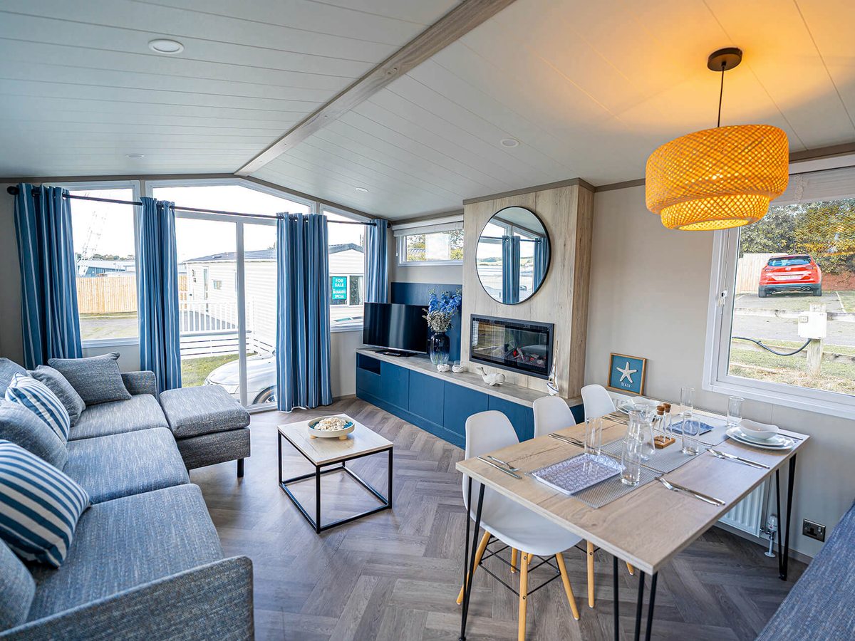 Bright open-plan living and dining room with a grey-blue corner sofa, a herringbone floor, a wide TV standing on a long blue media unit beside an inset fire and a round mirror, and a laid dining table for four.