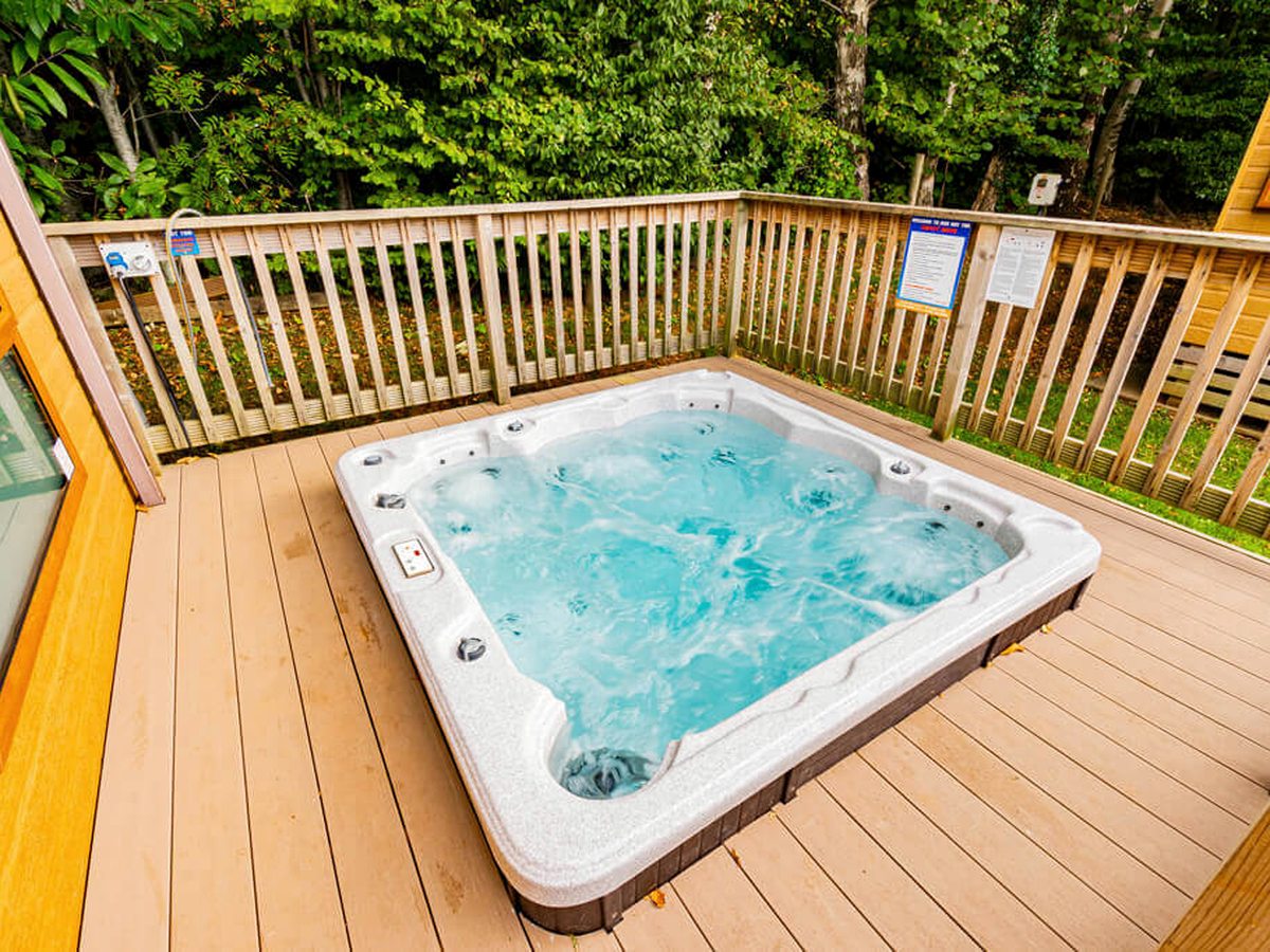 A square hot tub set into a timber deck outside a lodge, its water bubbling, with a wooden balustrade and trees behind.