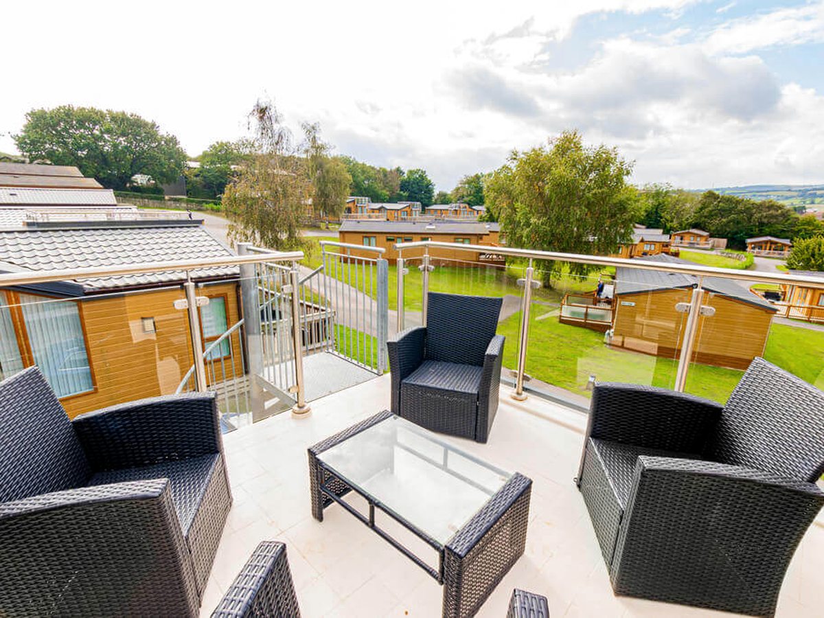 Rattan armchairs and a glass-topped table on a tiled first-floor veranda with a glass balustrade, above timber-clad lodges, mown grass and trees.